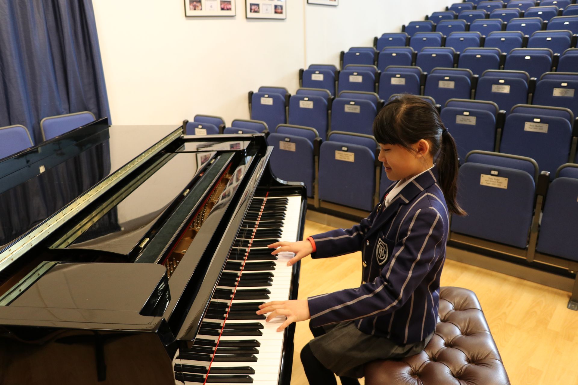 Girl in blazer playing grand piano in auditorium.
