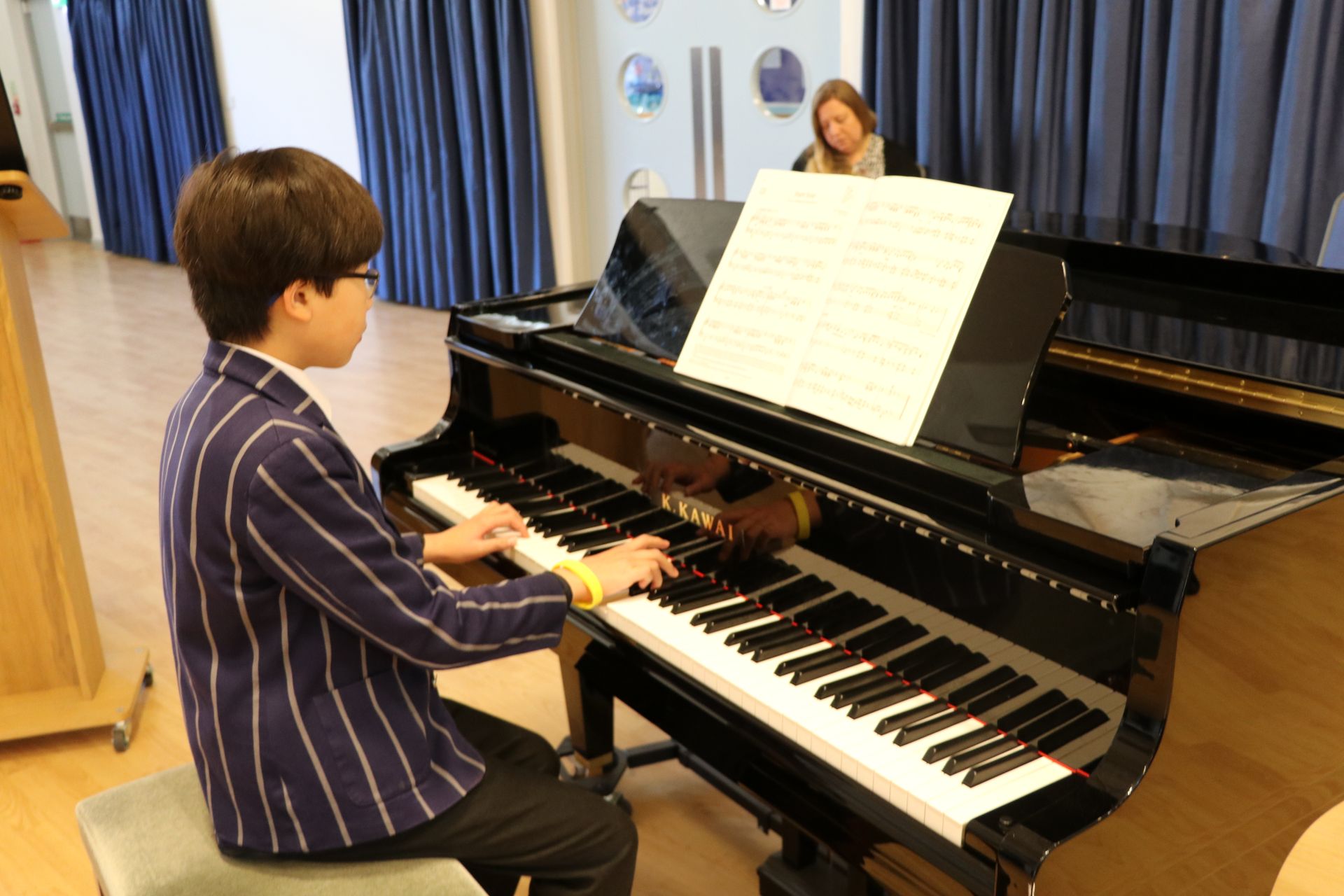Boy in school uniform plays piano, with sheet music. A woman observes from behind.