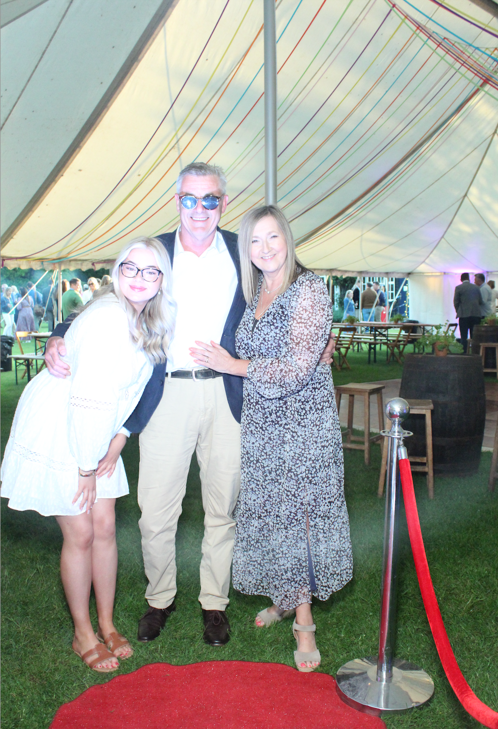 Family of three smiles under a tent on a red carpet. Man in sunglasses, woman in dress, and girl in white.