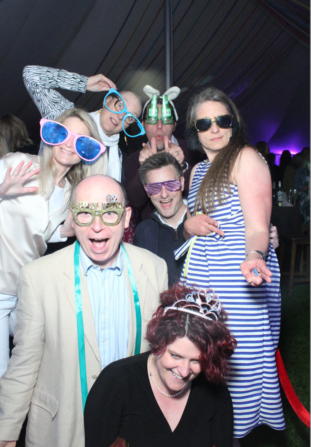 Group of people in silly glasses, posing and smiling at a party, indoors, some with arms raised, others smiling.