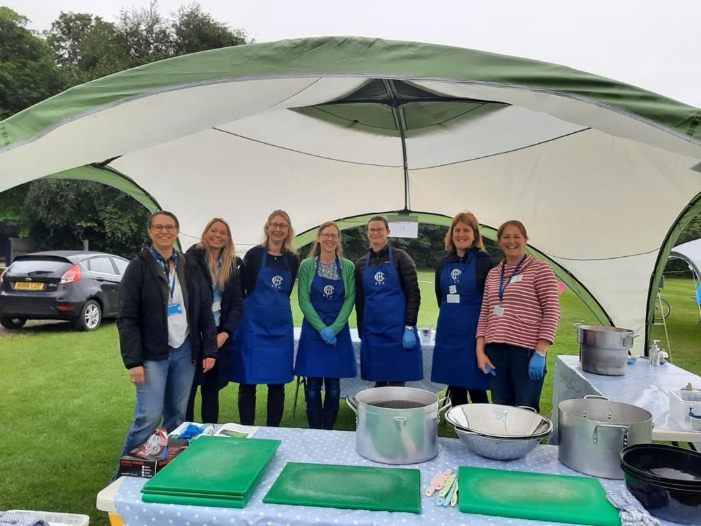 Seven people wearing aprons stand behind a food service table under a white tent.