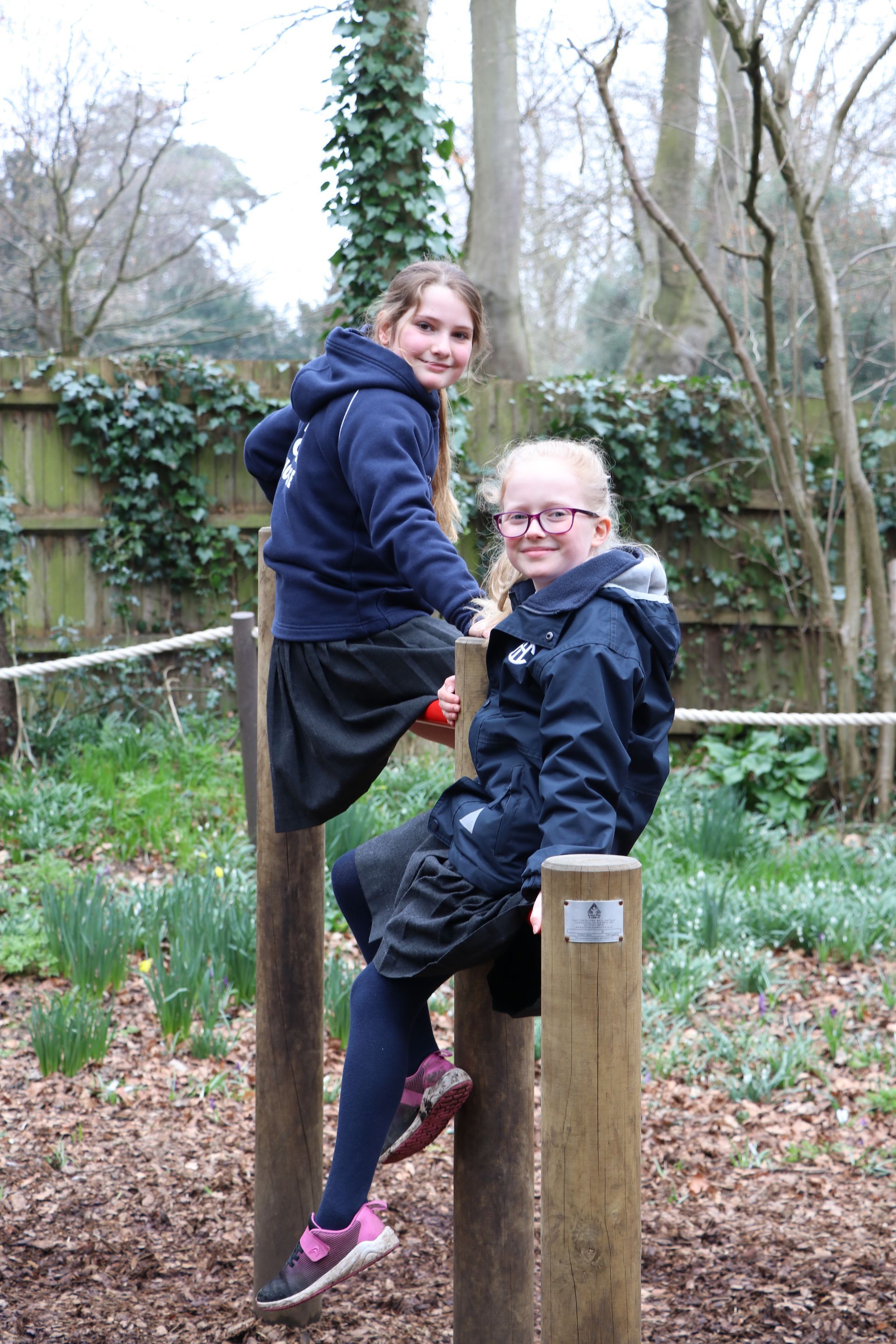 Two girls in blue jackets sit on wooden posts outdoors, smiling.