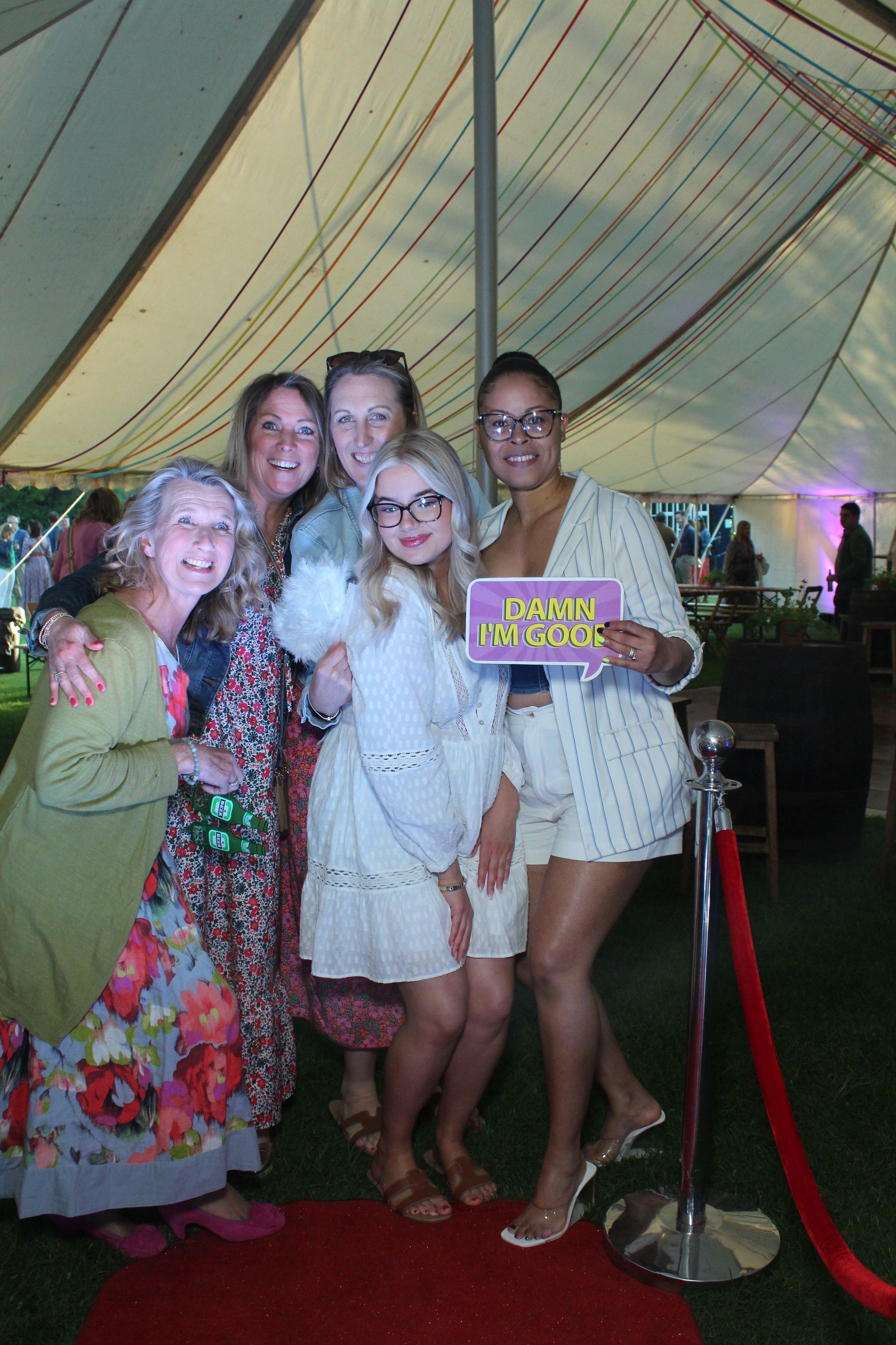 Five women smiling, posing on a red carpet at an outdoor event under a tent. One holds a 