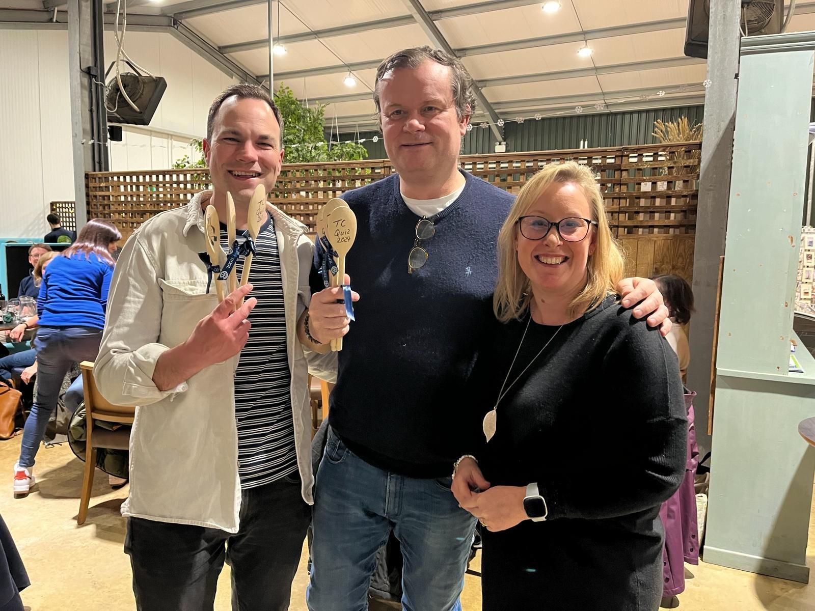 Three people smiling, holding wooden spoons, indoors. One in striped shirt, one in a sweater, one in black top; people in background.