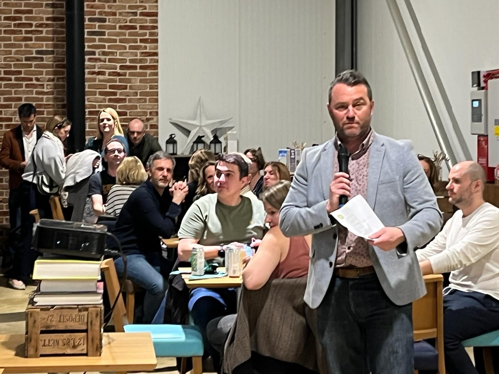 Man in a light blue jacket speaks into a microphone, addressing a seated audience in a brightly lit room.