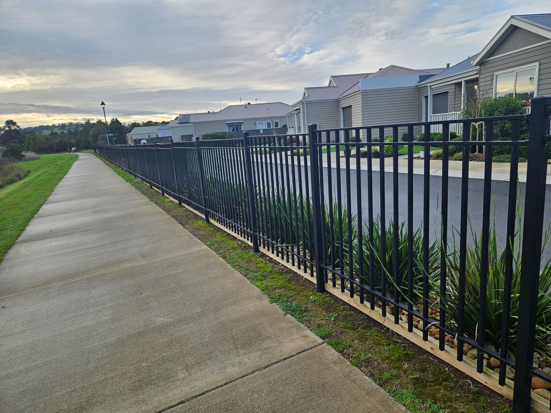 A fence along a sidewalk in a residential area with houses in the background.