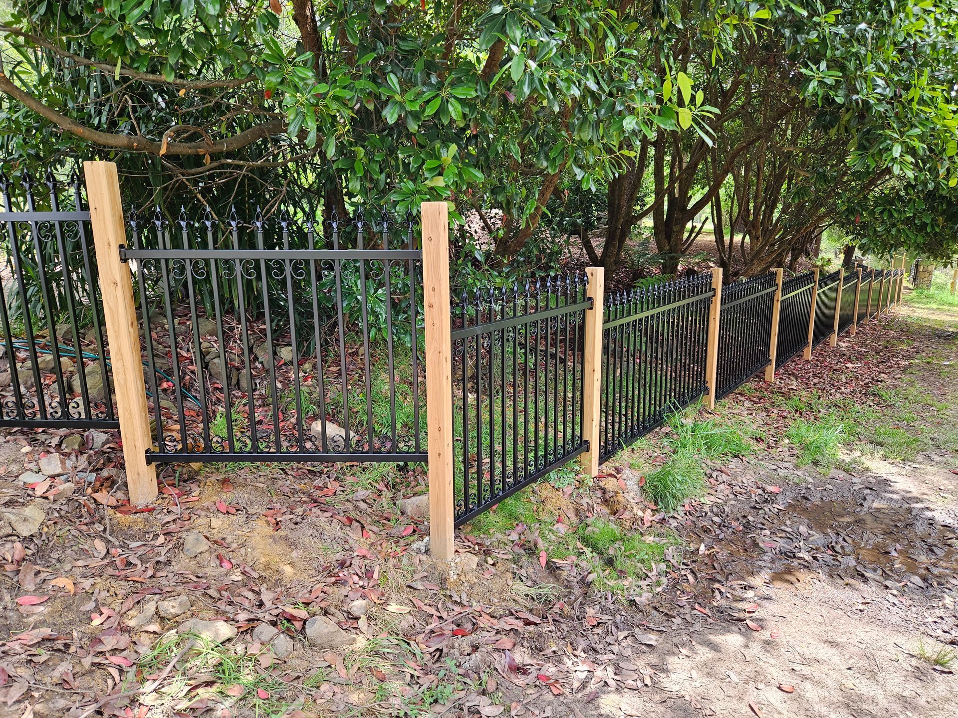 A black metal fence with wooden posts is surrounded by trees.