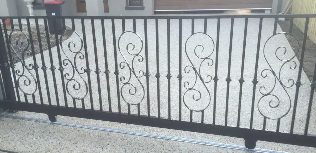A wrought iron gate with a swirl design is sitting on top of a gravel driveway.