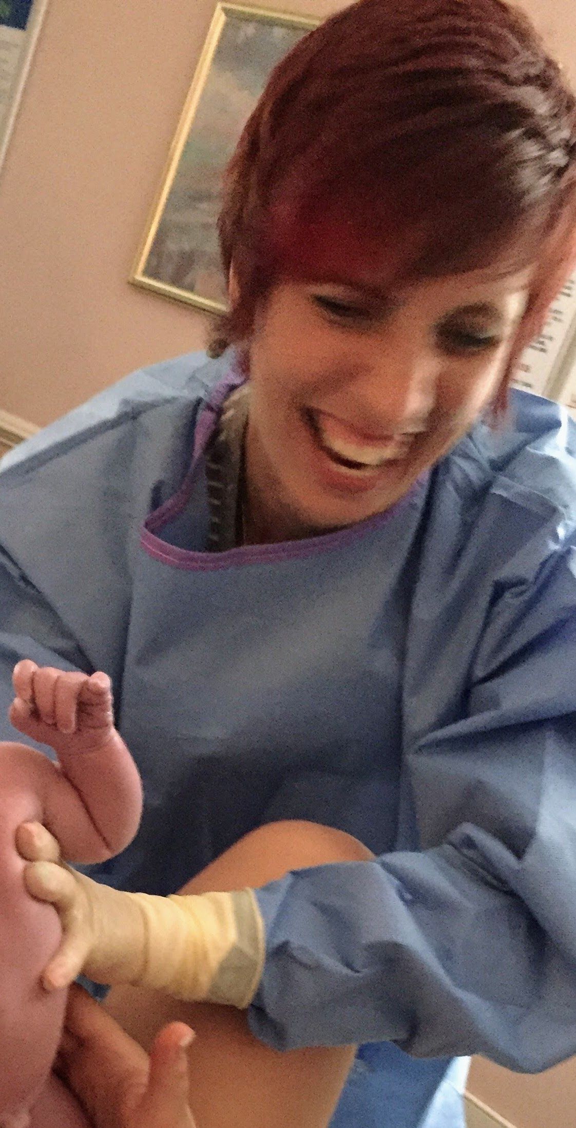 Midwife in a blue medical gown smiling at a new mother while holding a baby right after birth