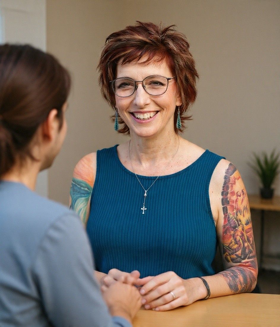 Two people in counseling at a table; smiling therapist with glasses and colorful arm tattoo faces the camera.