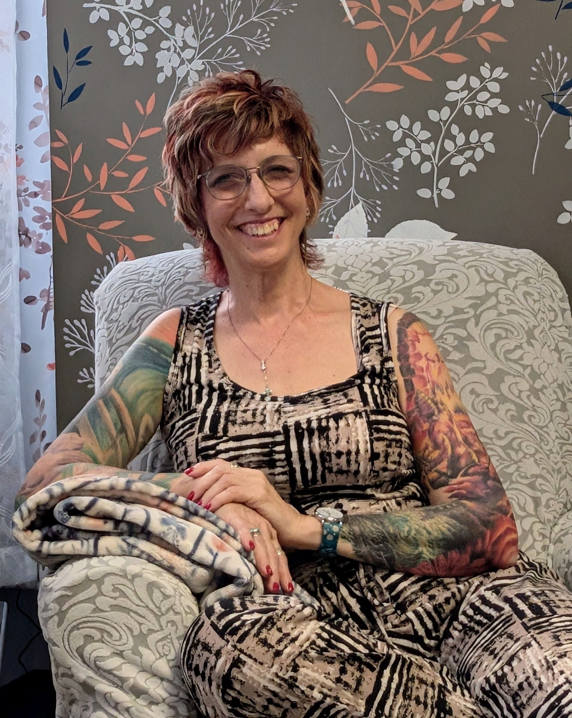 Smiling therapist with colorful tattoos seated on a patterned chair against floral wallpaper.