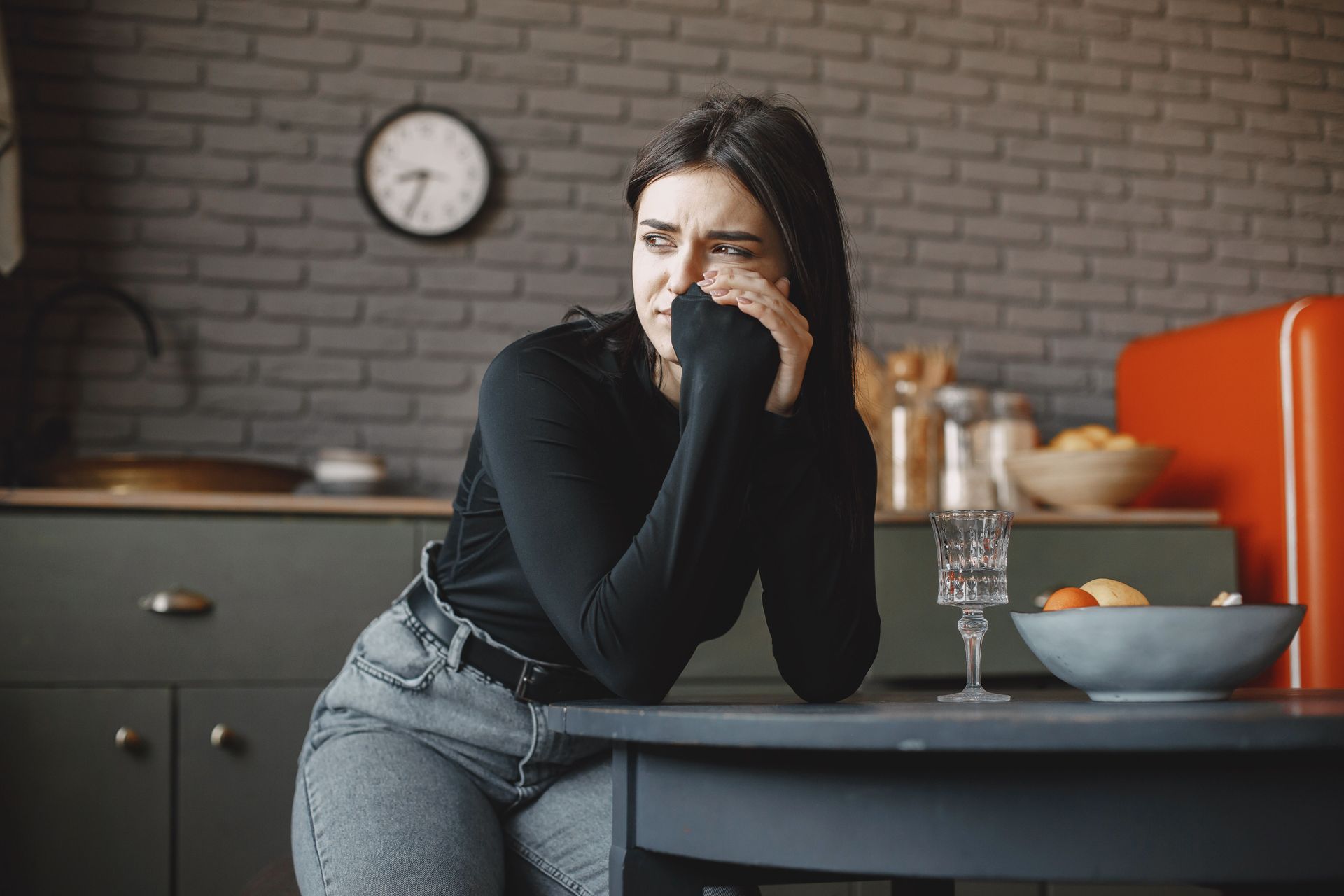 Woman with PTSD sits at a table, looking pensive about pregnancy or motherhood in a kitchen with a bowl and glass nearby.