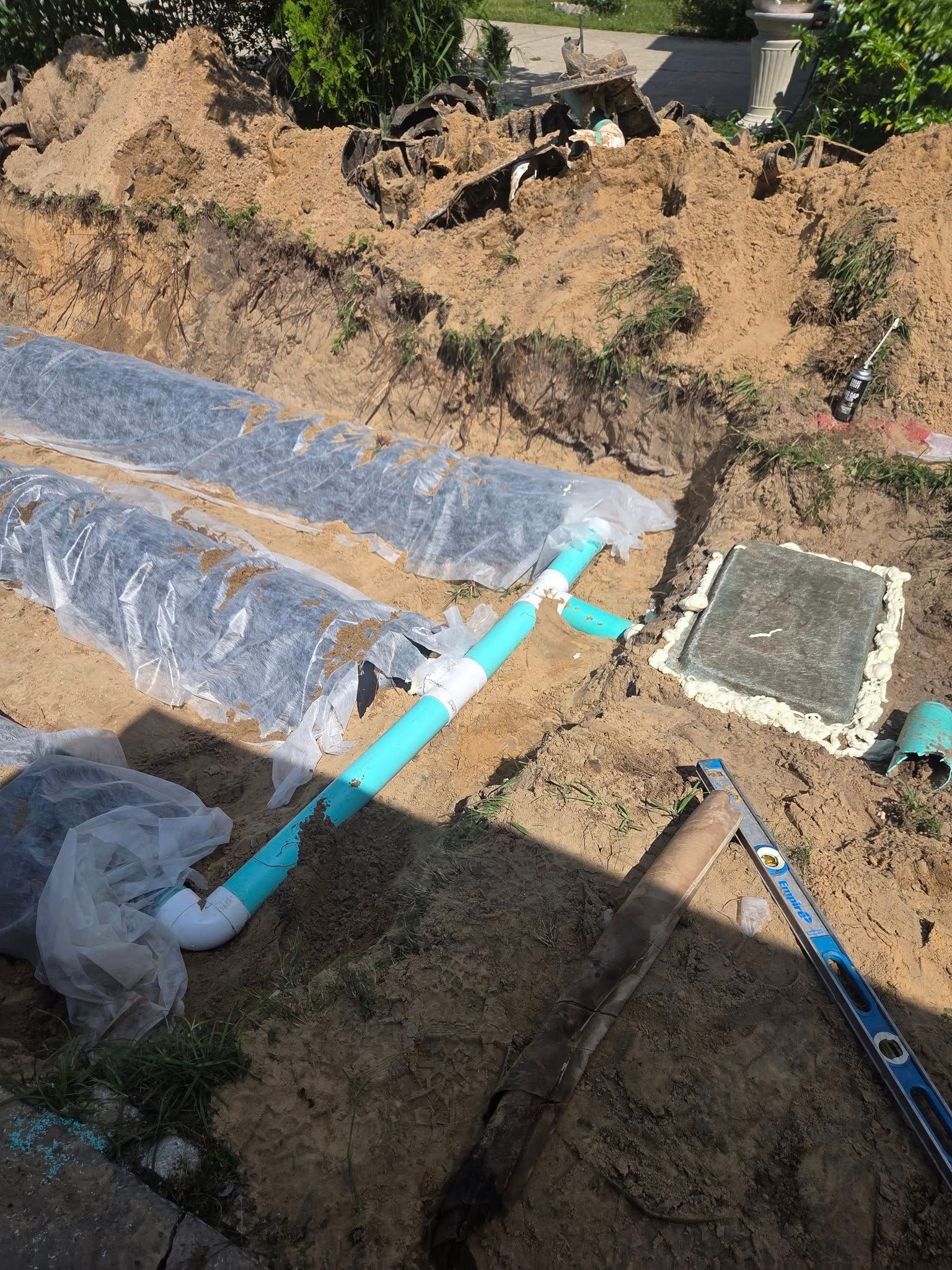 Underground septic system construction; blue pipes, gravel beds, plastic sheeting, and a concrete access cover in a dirt trench.
