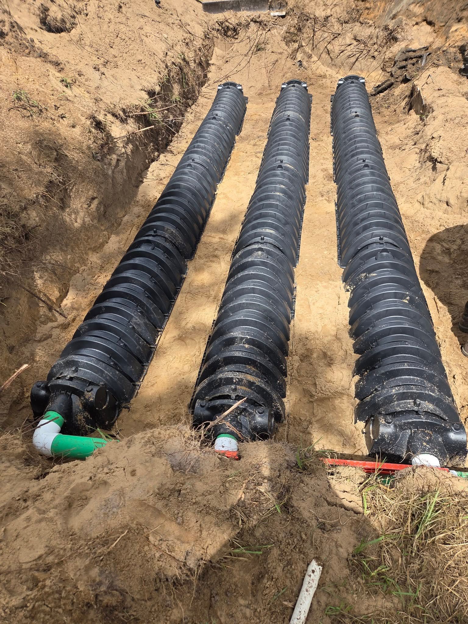 Three corrugated black pipes installed in a trench. Green and red pipes are connected, with dirt surrounding.