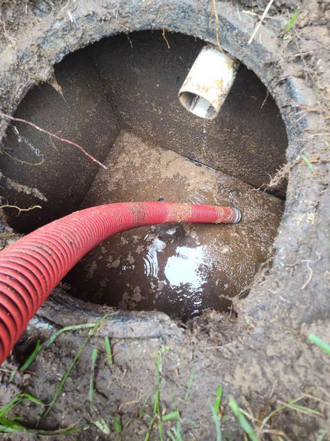 A septic tank with a red hose inside, surrounded by dirt and grass. A white pipe is visible.