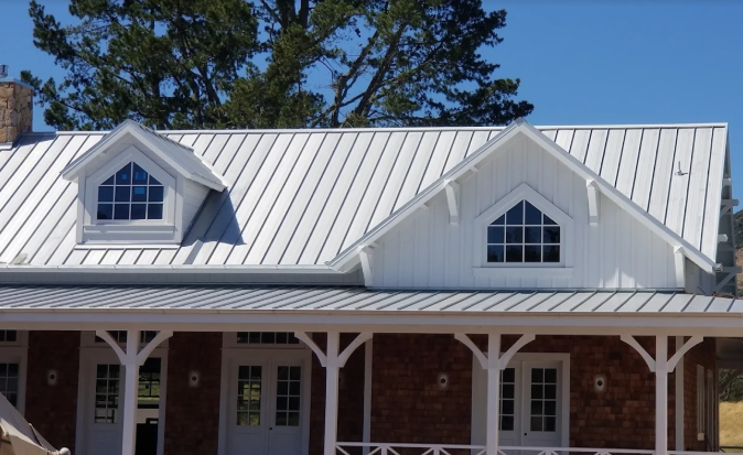 White house with a metal roof, two dormers, and a brick facade, against a blue sky.
