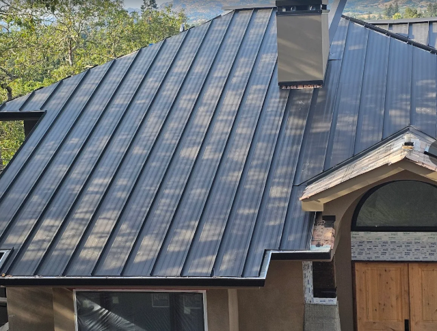 Black metal roof on a house with a chimney and part of a wooden facade in sunlight.