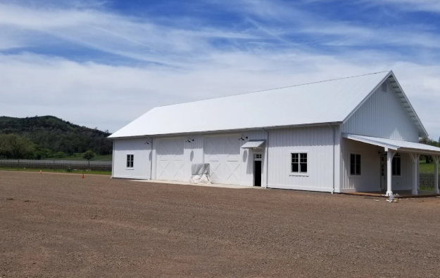 White barn-style building with a covered entrance, set in a gravel lot with a blue sky background.