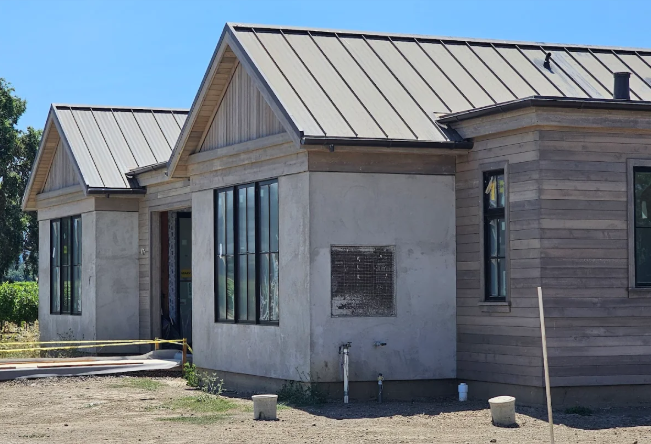 A modern house under construction with a metal roof and large black-framed windows, against a blue sky.