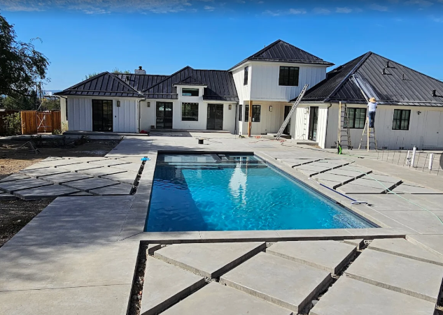House with a pool under construction; white exterior, black roof, blue pool water, concrete patio.