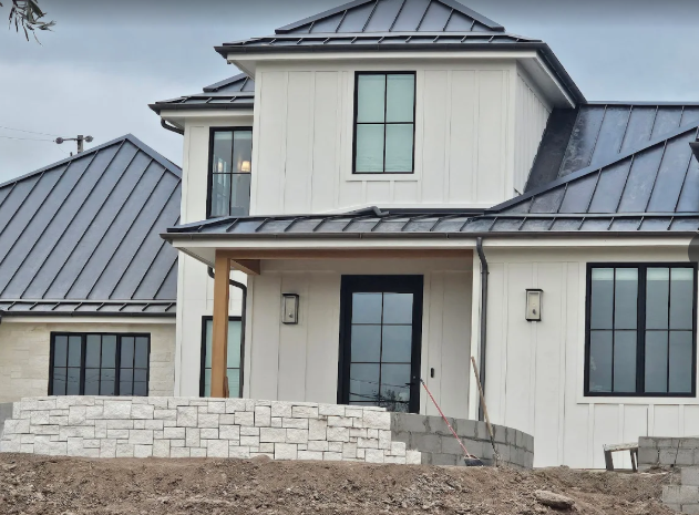 White farmhouse with black window frames, gray metal roof, stone foundation, overcast sky.