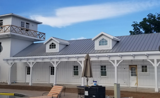 White building with gray metal roof, dormers, porch, and tower against a blue sky.