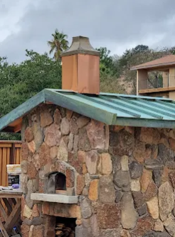 Stone oven with copper chimney on a green metal roof.