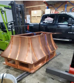 Copper chimney cap on a pallet, ready for transport in a warehouse; a forklift and truck are in the background.