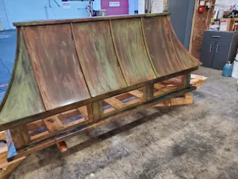 Copper hood with patina finish, resting on a wooden pallet in a workshop.