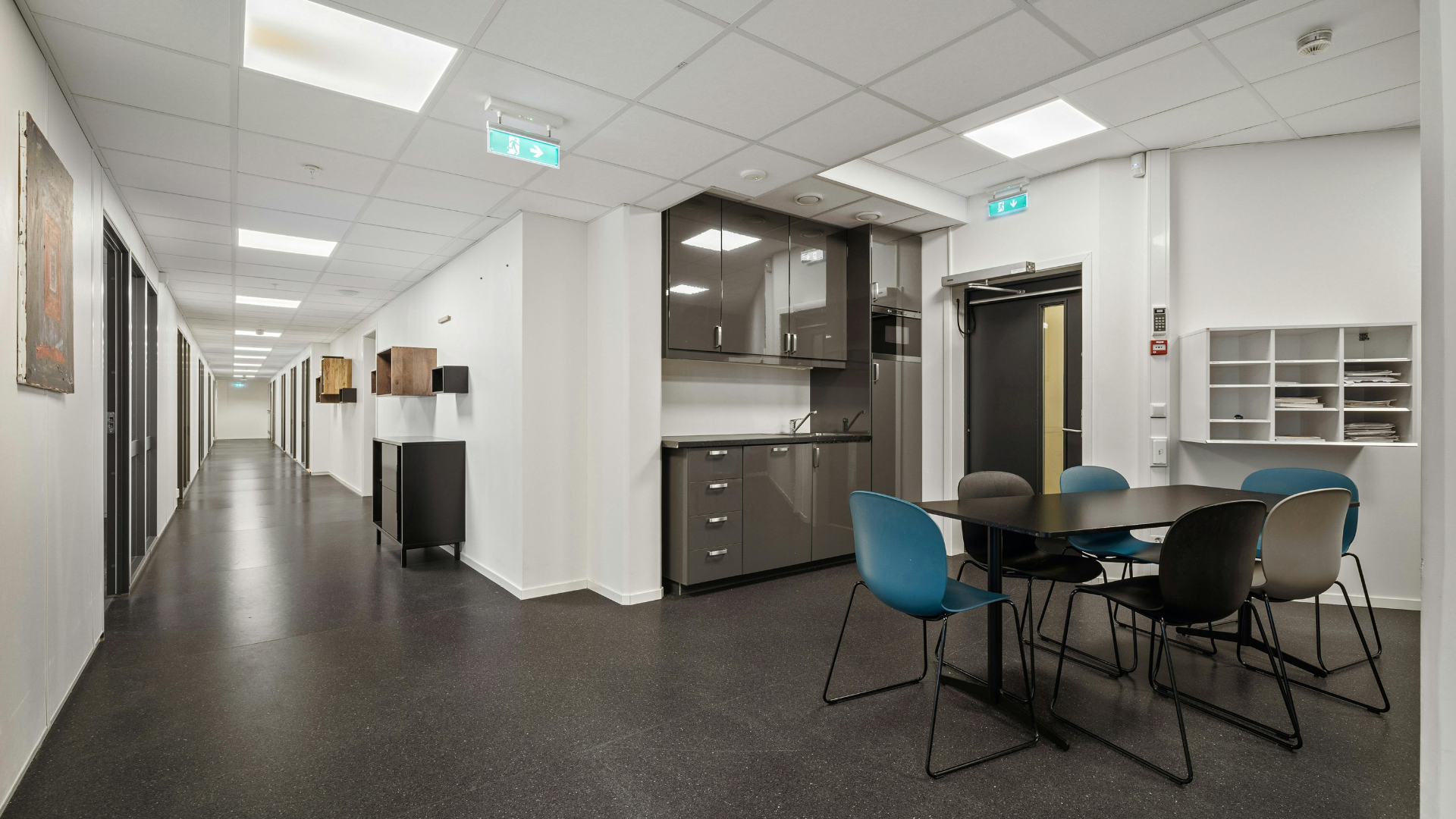 Long hallway with a kitchen area and a table with chairs. Dark flooring, white walls, and several doors along the hall.
