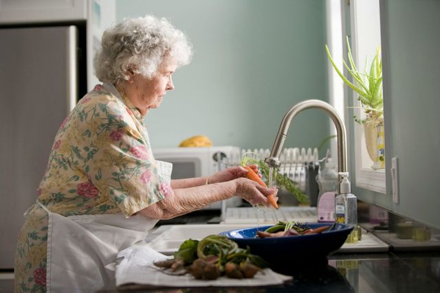 An elderly woman is washing vegetables in a kitchen sink.