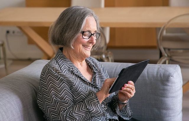 An elderly woman is sitting on a couch using a tablet computer.