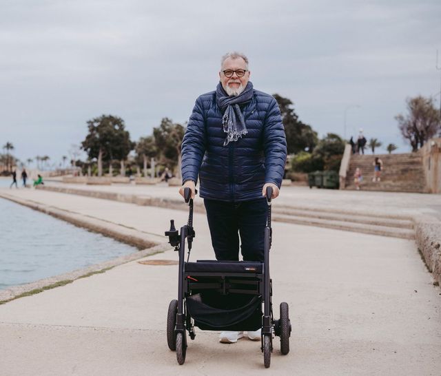 An elderly man is pushing a walker on a sidewalk next to a body of water.