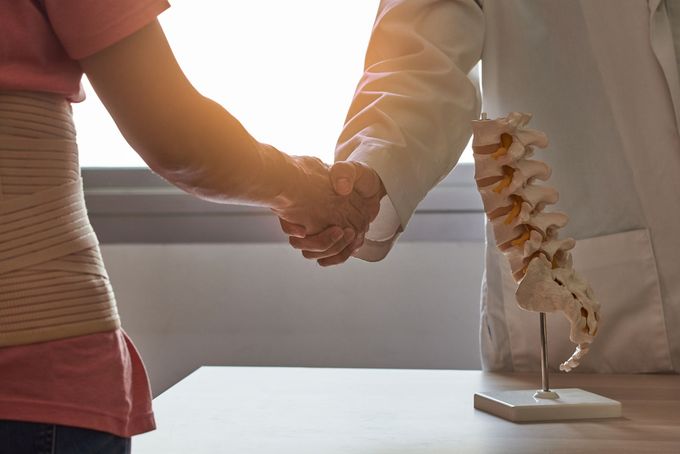 Un paziente con un tutore per la schiena stringe la mano a un medico vicino a un modello della colonna vertebrale.