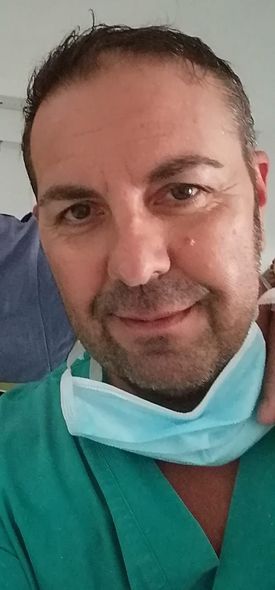 Uomo che indossa un camice medico e tiene in mano una mascherina. Ha i capelli scuri e sorride leggermente.