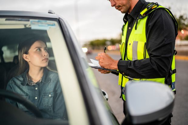 A police officer in a yellow reflective vest writes on a clipboard while standing by a car with a driver inside.