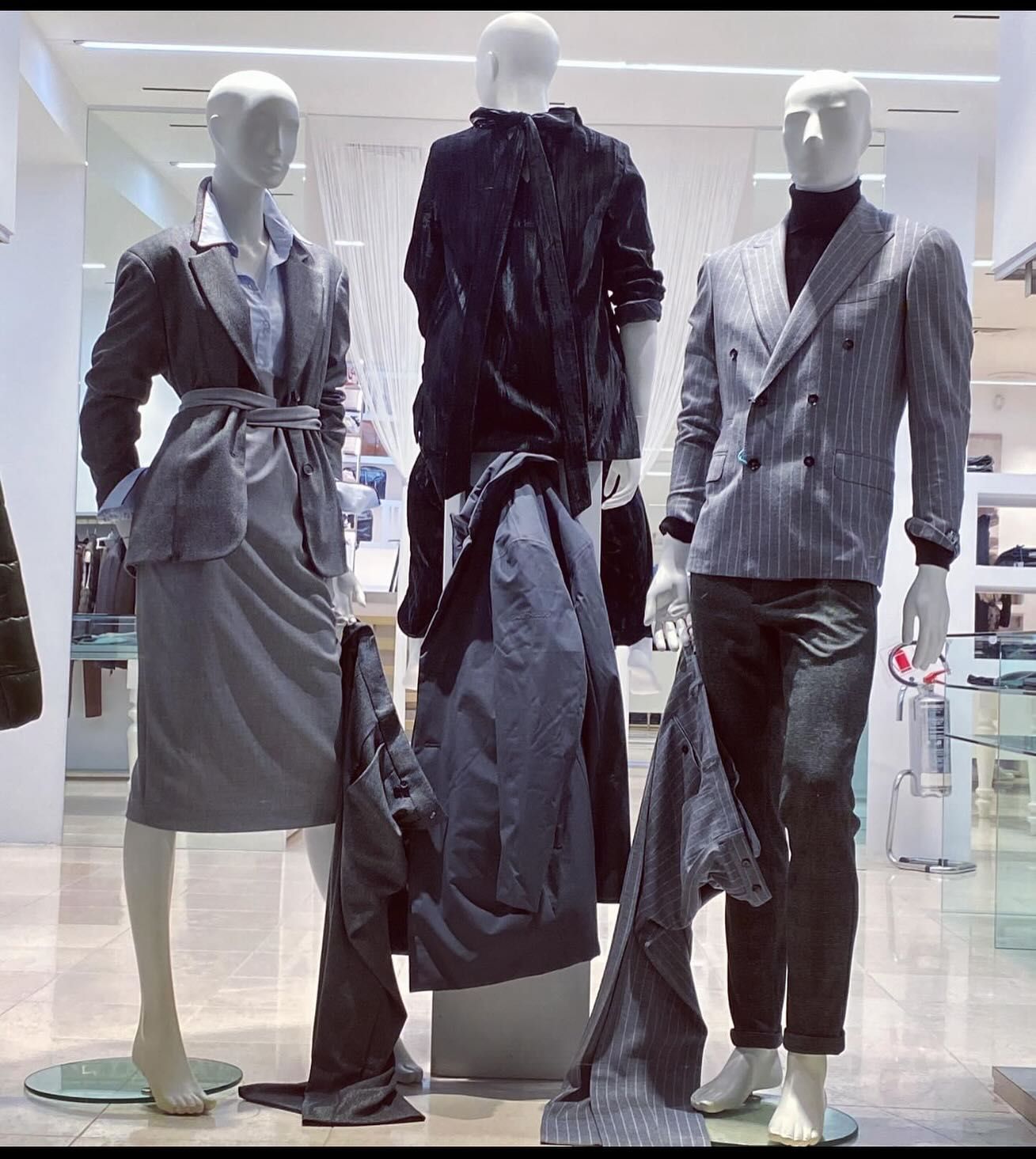 Tre manichini esposti in un negozio di abbigliamento: una donna in abito grigio e due uomini in giacca e pantaloni.