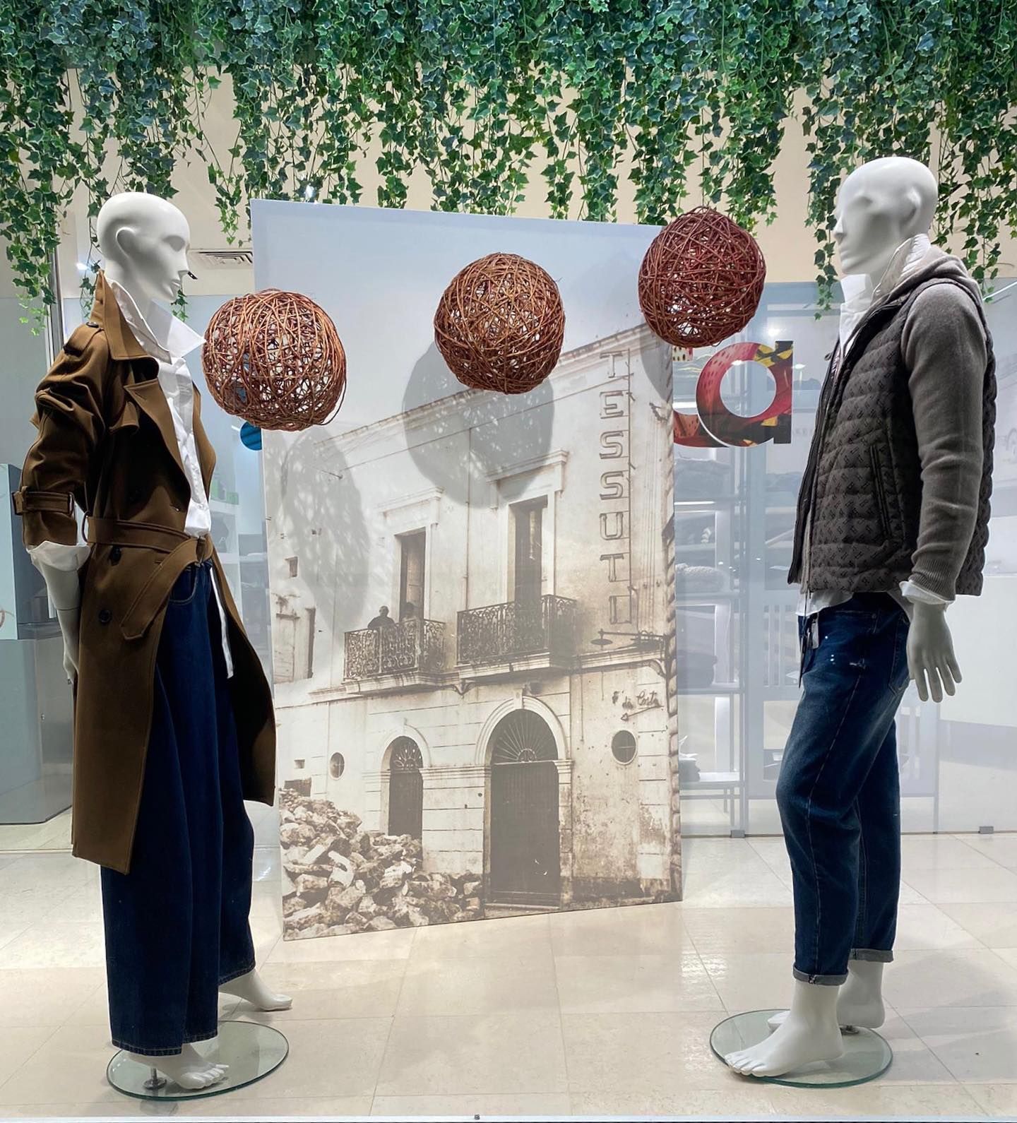 Due manichini in una vetrina, uno con un cappotto marrone, l'altro con un piumino, davanti a un edificio sullo sfondo.