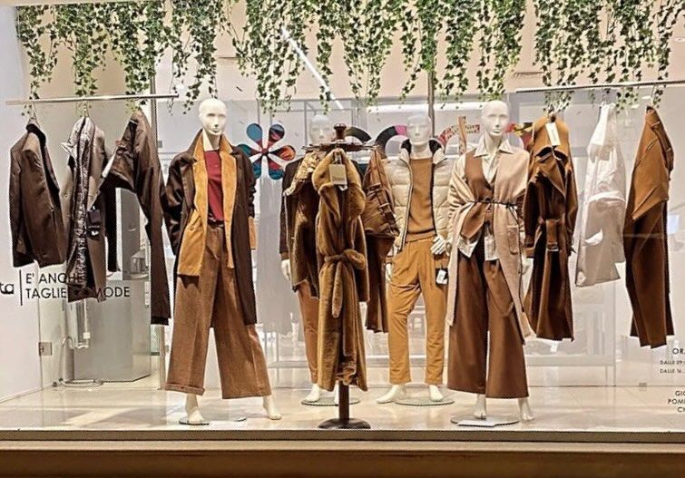 Vetrina con manichini vestiti di marrone e beige, piante in alto.