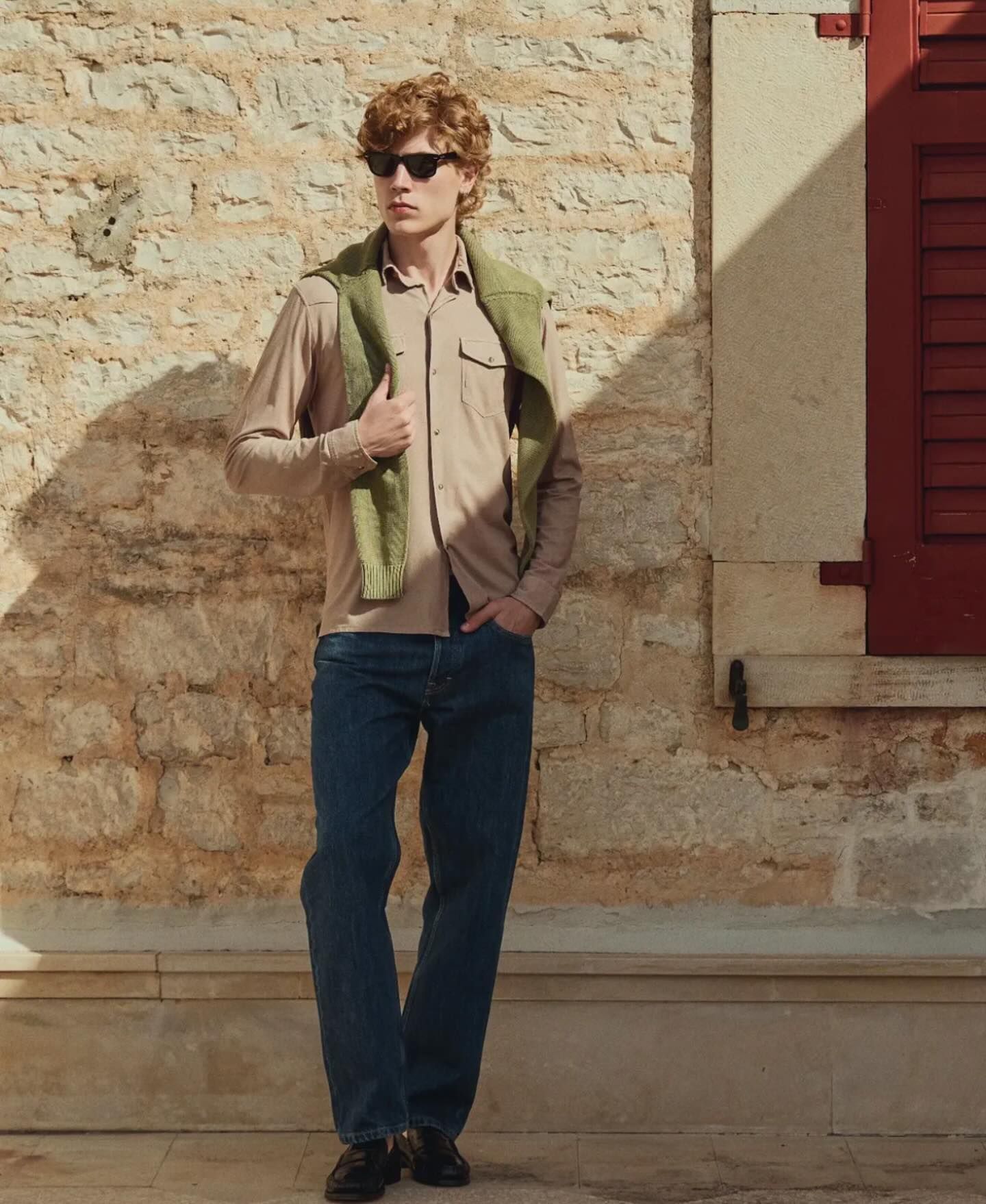 Un uomo con occhiali da sole, camicia e maglione al collo è in piedi contro un muro di pietra.