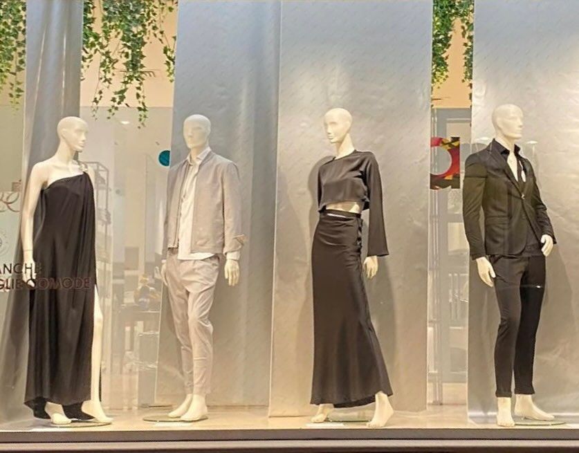 Vetrina con quattro manichini in abiti dai toni neutri, in piedi davanti a pannelli trasparenti.