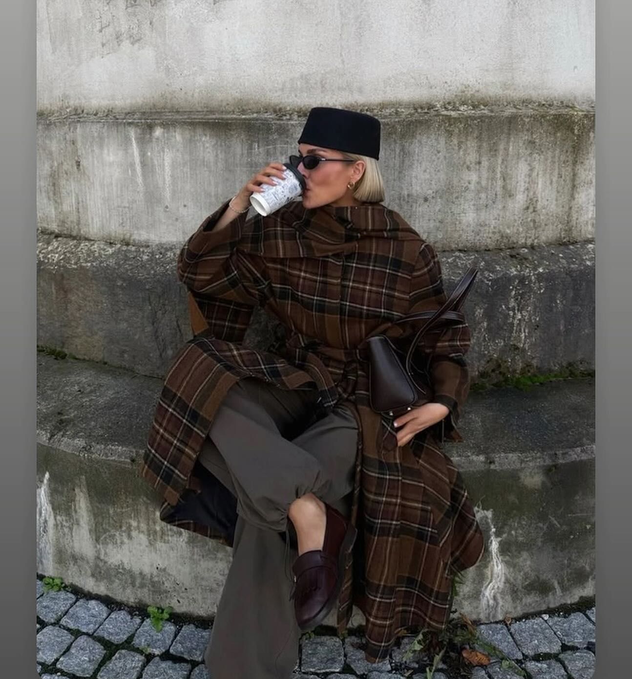 Una persona sorseggia un caffè da una tazza mentre indossa un cappotto scozzese, seduta all'aperto.