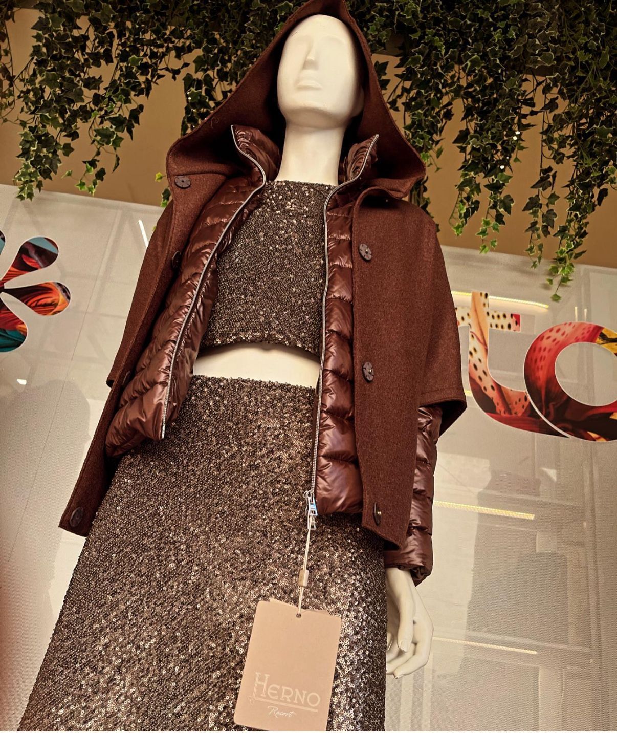 Persona con giacca con cappuccio color marrone e pantaloni sopra una camicia bianca a maniche lunghe, in posa su uno sfondo bianco.