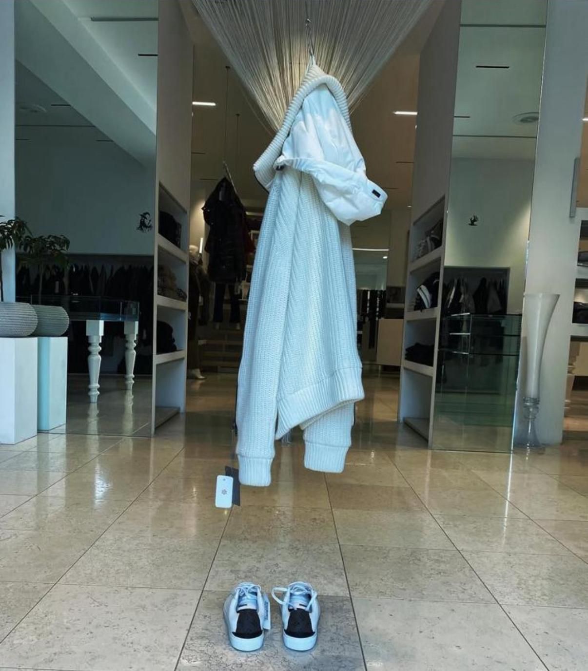 Manichino in un negozio che indossa un maglione marrone chiaro, pantaloni marroni e una sciarpa a quadri, in piedi vicino a scaffali pieni di vestiti piegati.