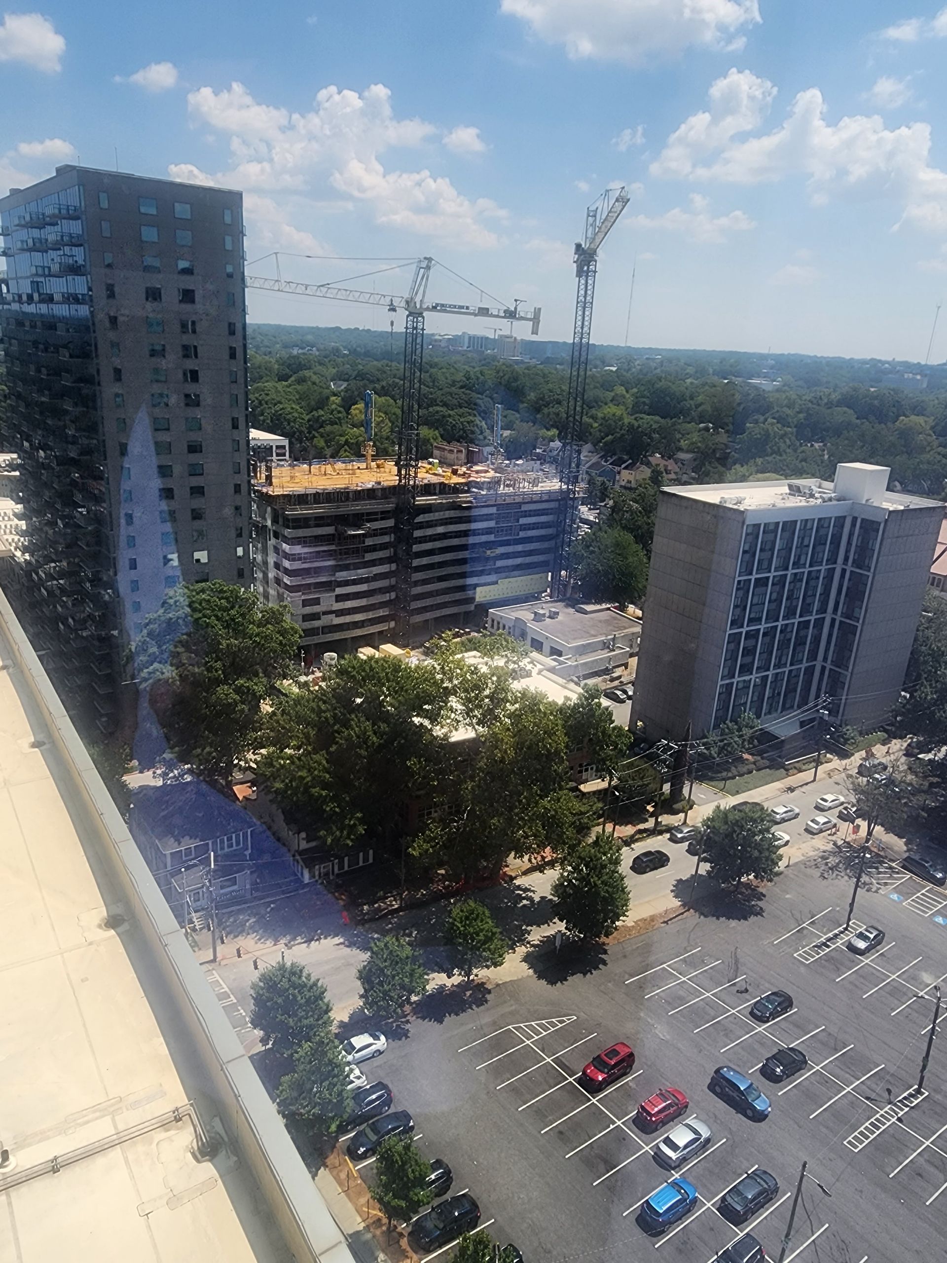 Skyscrapers and construction site with cranes amidst trees and parking lot under a blue sky.
