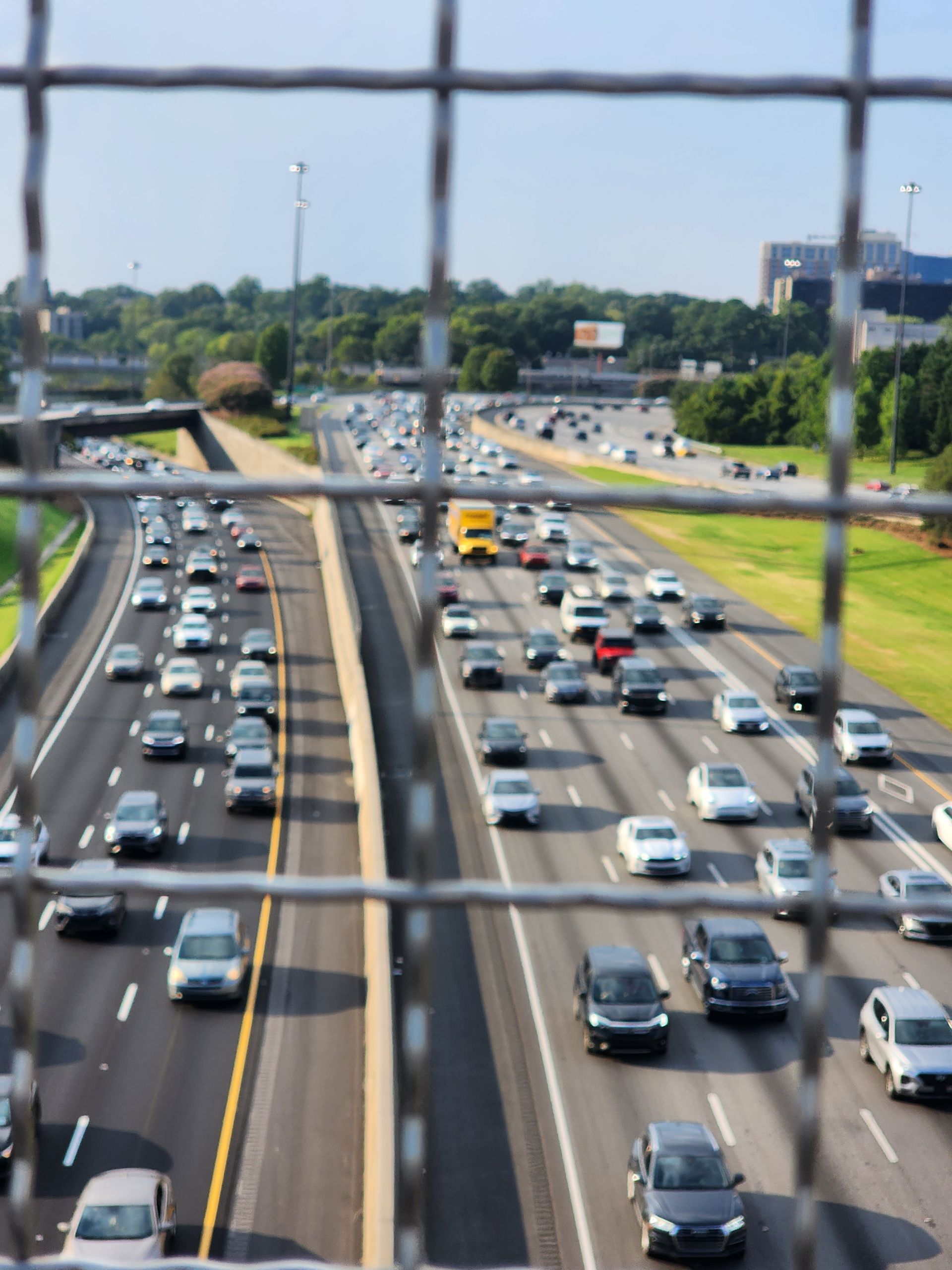 Highway traffic viewed through a metal fence. Cars move along multi-lane roads. Green trees and blue sky visible.