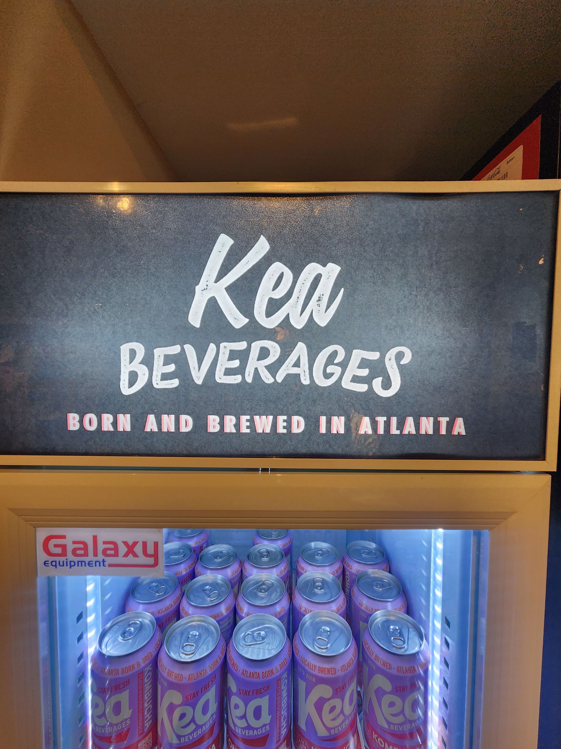 Mini refrigerator displaying Kea Beverages cans. Sign reads 