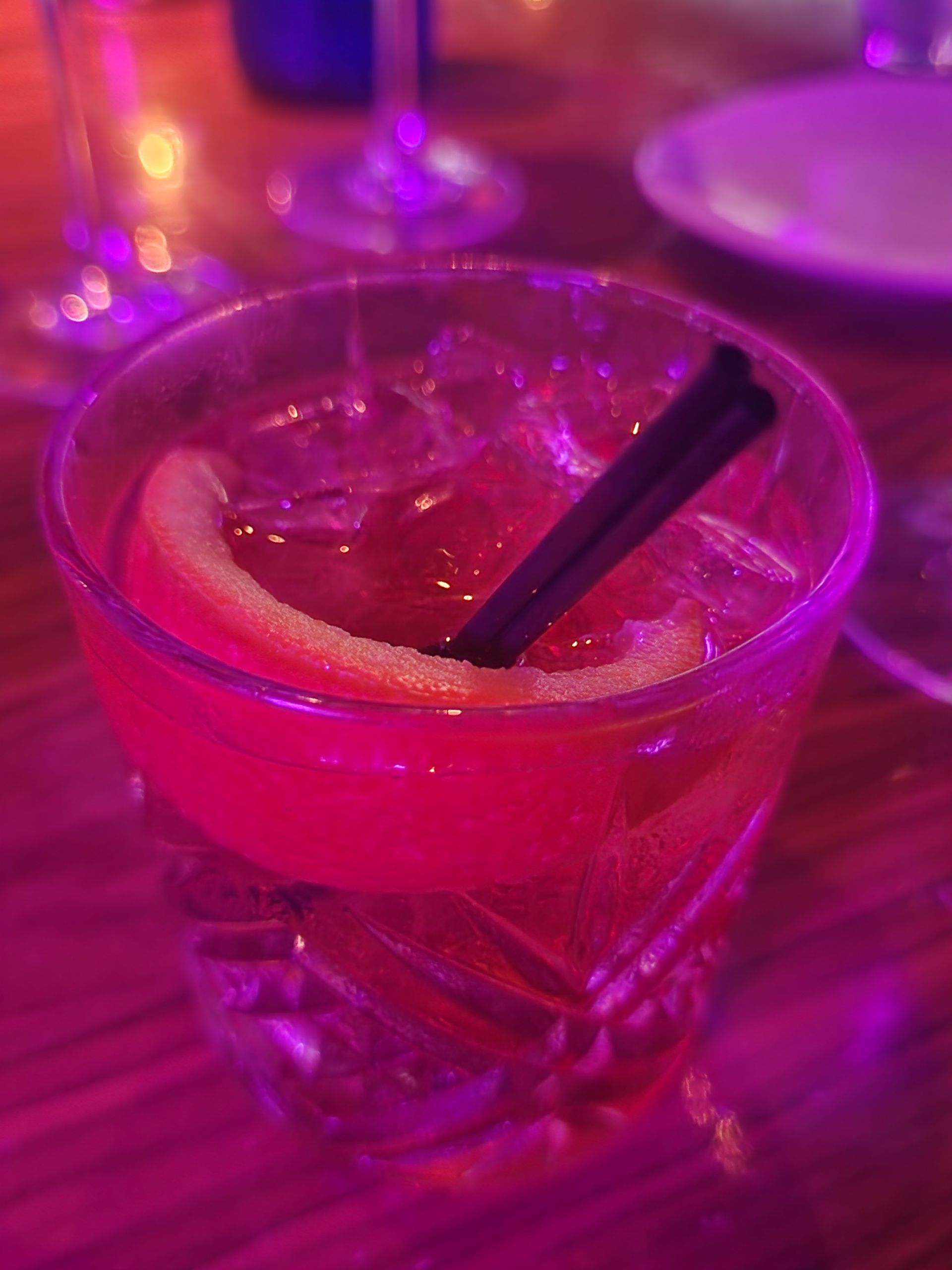 Cocktail with orange slice and black straw in a patterned glass, lit with purple light.