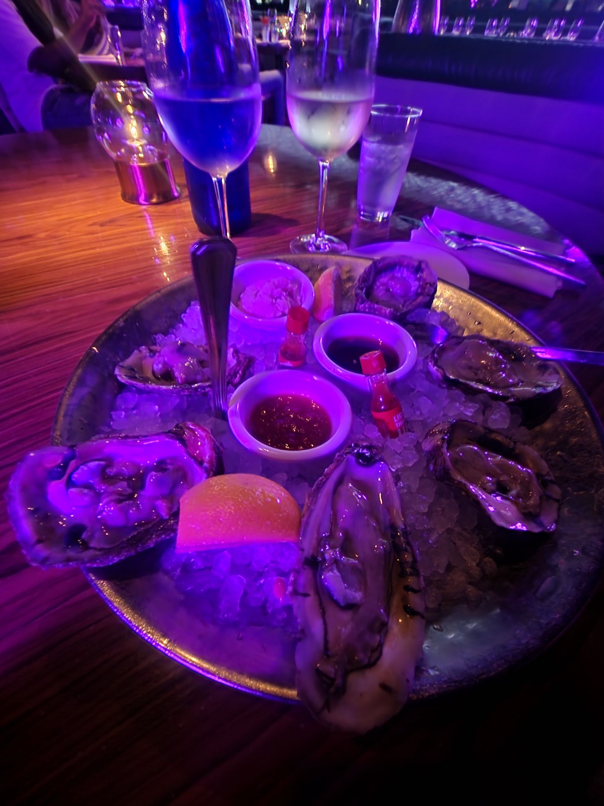 Oyster platter with sauces, lemon, and glasses of wine on a table, with purple lighting.