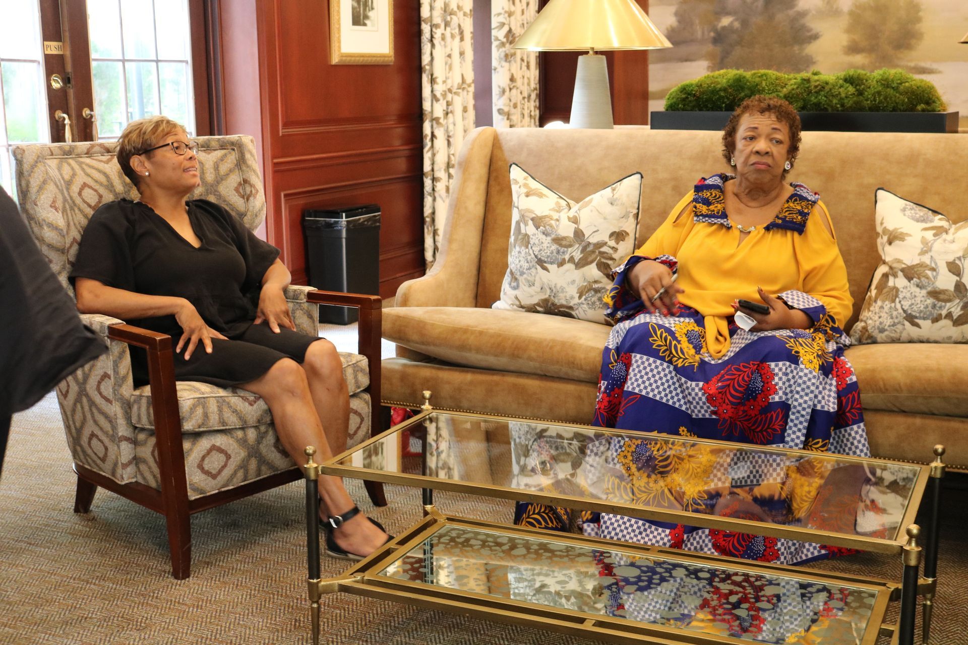 Two women seated indoors: one in black, the other in yellow and patterned fabric. They are in a living room setting.